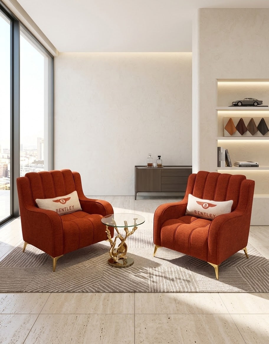 Two matching orange armchairs with gold legs and white pillows in a modern, sunlit living room. A small round glass-top side table sits between them on a gray patterned rug.